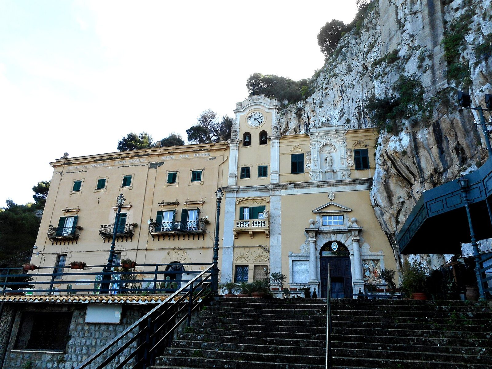 La facciata gialla del Santuario di Santa Rosalia con il campanile, addossata alla parete di roccia del Monte Pellegrino, e la scalinata d'accesso