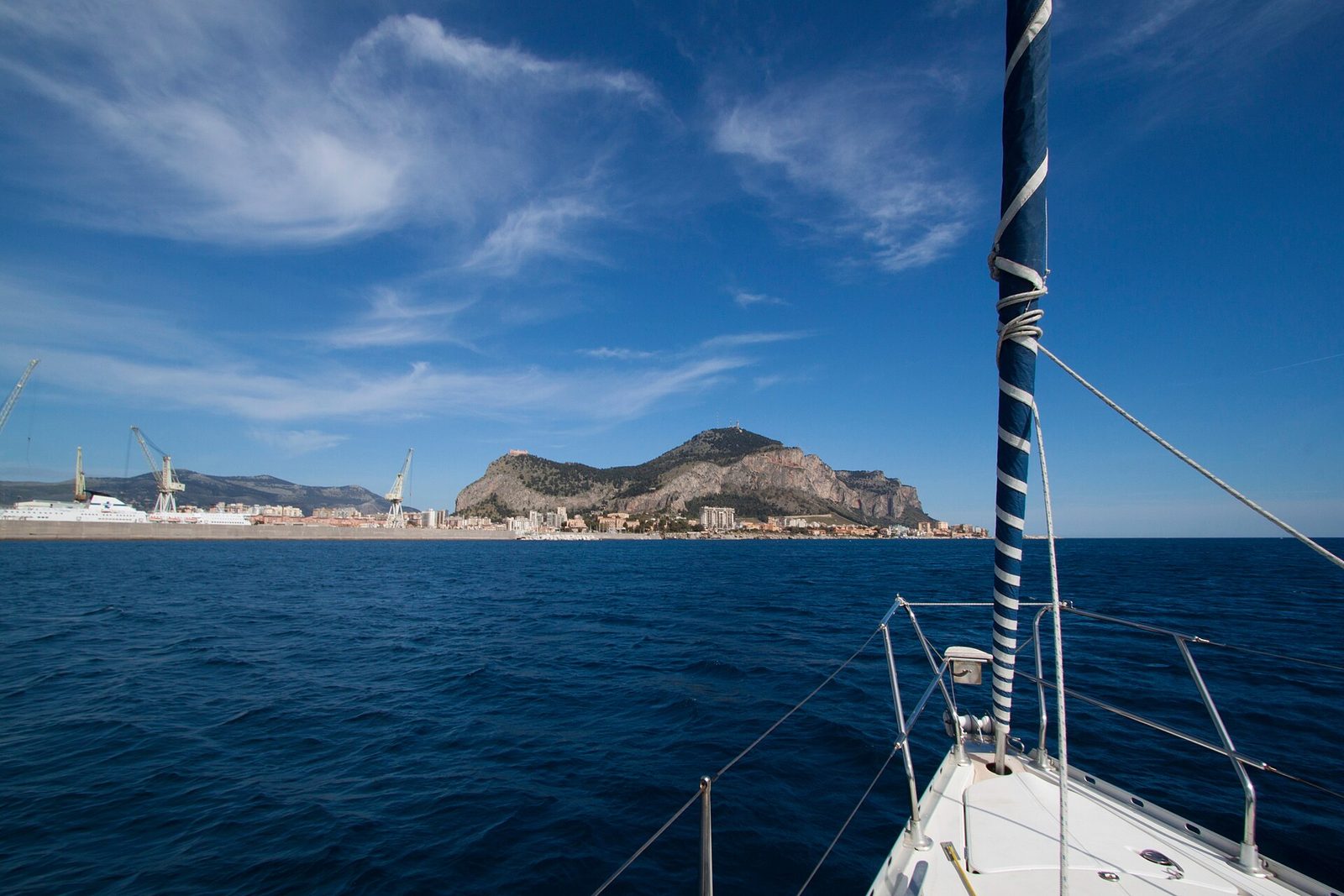 Il golfo di Palermo visto dalla prua di una barca a vela, con il mare blu, il porto e il Monte Pellegrino sotto un cielo con nuvole leggere